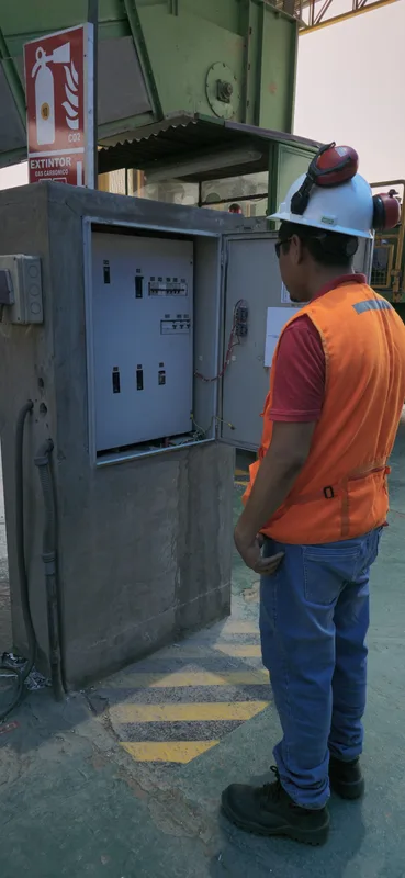 Técnico con casco, lentes y chaleco revisando tablero en cuarto con extintor CO2 y señalética